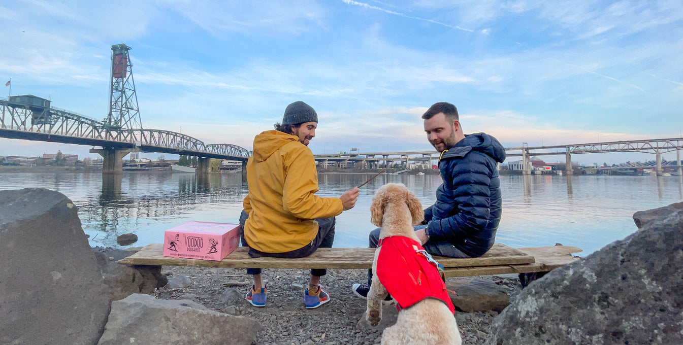 Two men sit by the water in a city with their dog.