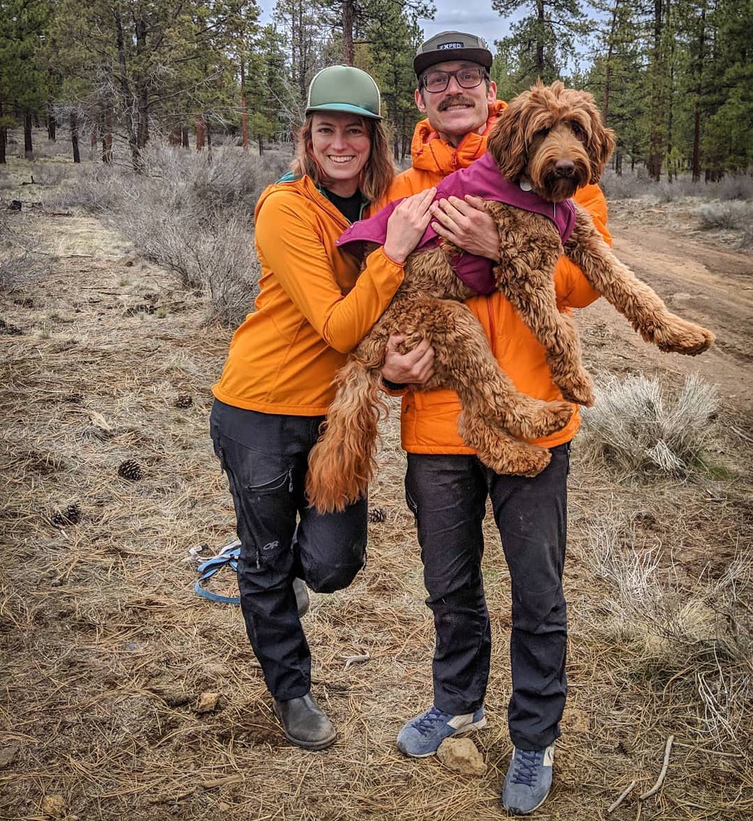 graham and shannon in matching jackets and pants hold up pebble while standing next to a forest road outside.