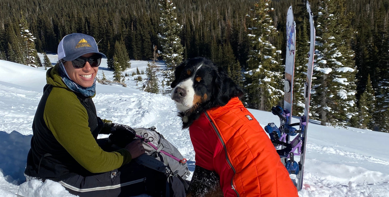 A woman wearing skis sits on a snow-covered mountain with her Bernese Mountain Dog. 
