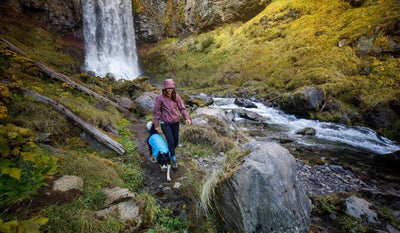 Woman walks along river and waterfall with dog in wind sprinter jacket.