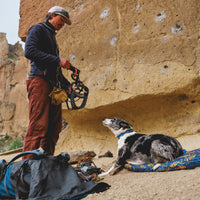 A man puts on a rock climbing harness with his dog who's on a bed.