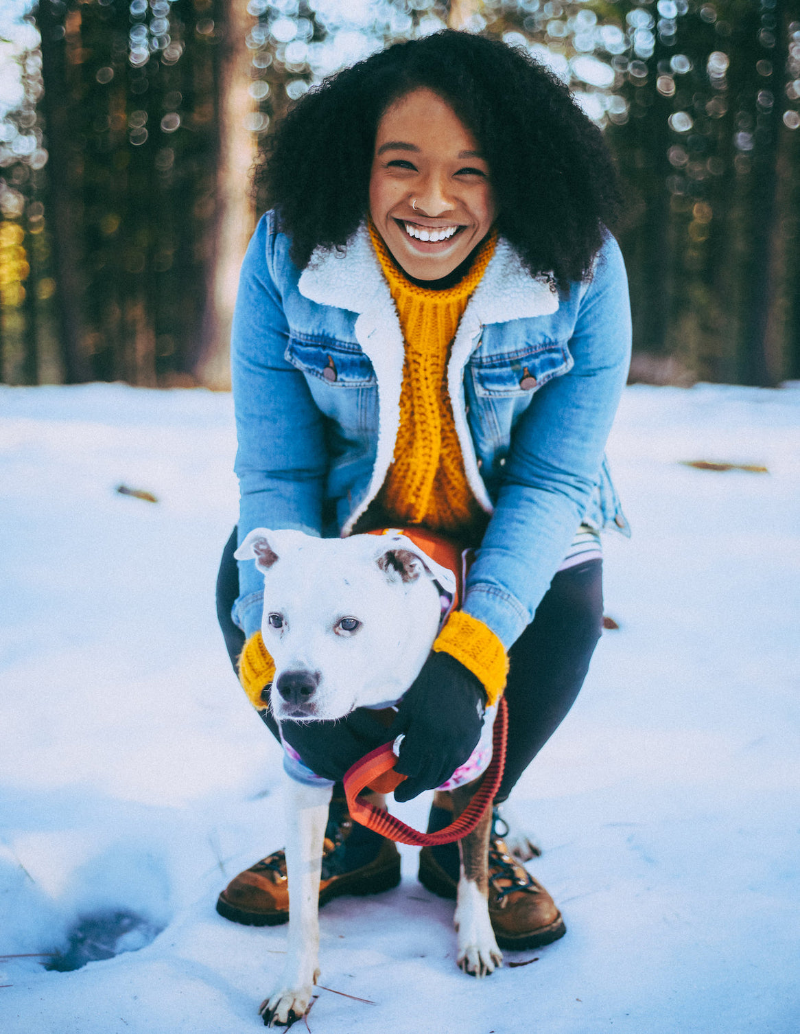 Alexis in jean jacket poses with her white pitbull mix on the snow in the woods. 