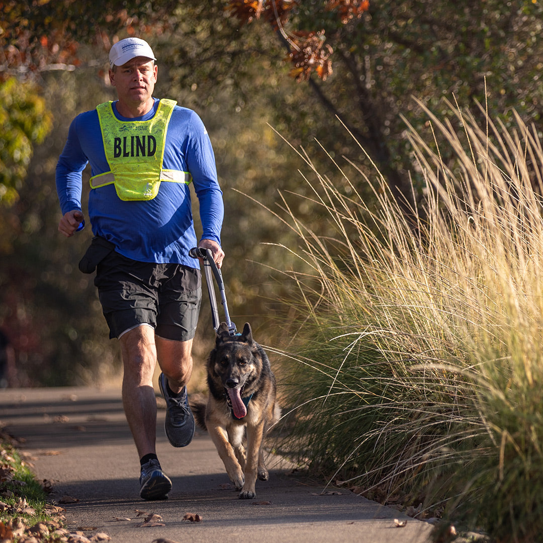 Athlete in blind vest runs with german shepherd guide dog in unifly harness on paved path.