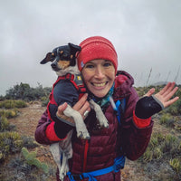 Katie smiles and holds her small dog in her arms mid hike.